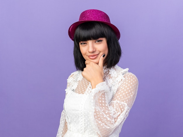 Thoughtful young party girl wearing party hat standing in profile view looking at front keeping hand on chin isolated on purple wall with copy space