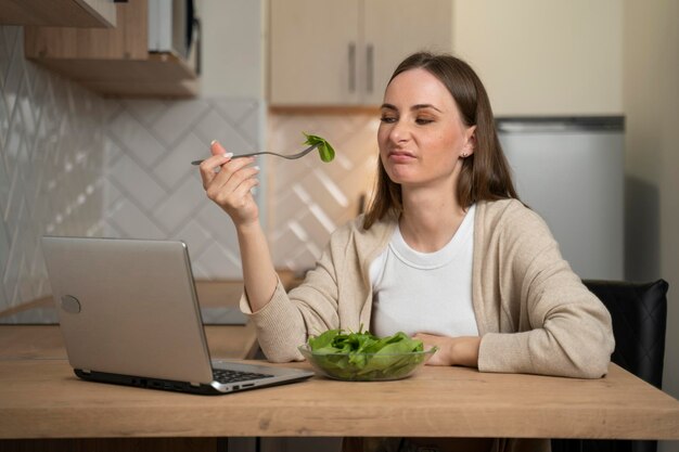 A thoughtful young woman eats a salad and experiences a lack of appetite while sitting in the kitchen and using a laptop Digestive problems as well as spoiled and tasteless food