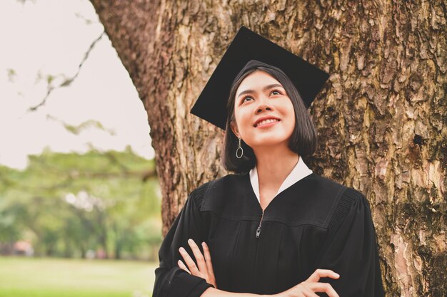 Photo thoughtful young woman in graduation gown with arms crossed standing by tree at park