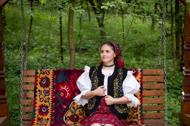 Фото Вдумчивая молодая женщина в румынской одежде сидит на качелях во дворе