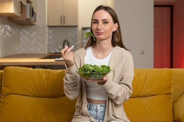 A thoughtful young woman is eating a salad and experiencing a lack of appetite while sitting on the couch Digestive problems as well as spoiled and tasteless food