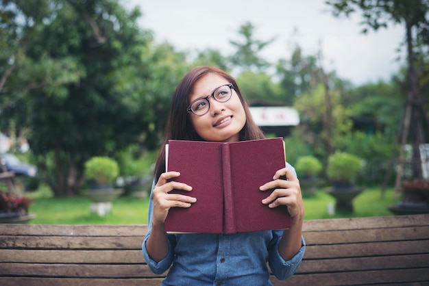 Foto giovane donna pensierosa che guarda lontano mentre tiene in mano un libro su una panchina del parco