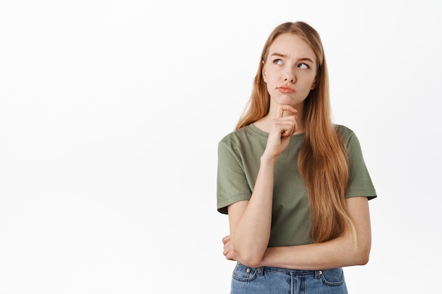 Thoughtful young woman making serious decision, looking up while pondering choices, frowning while thinking, standing in t-shirt and jeans against white wall