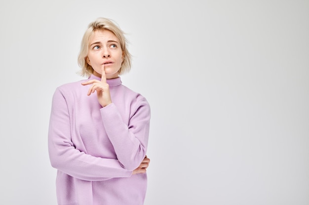 Thoughtful young woman in purple sweater with finger on her chin isolated on a light background