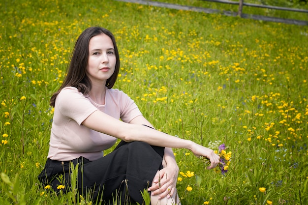 Thoughtful young woman sits on a flower meadow. Summer sunny day