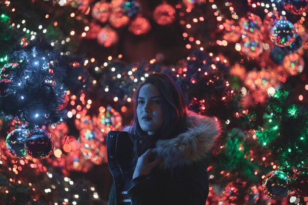 Фото Вдумчивая молодая женщина стоит на освещенной рождественской елке ночью