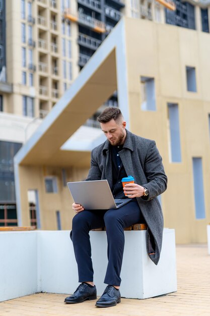 Thoughtfull businessman sitting with laptop and coffee on modern street. Clever man working on distance. Adult guy doing his work in internet.