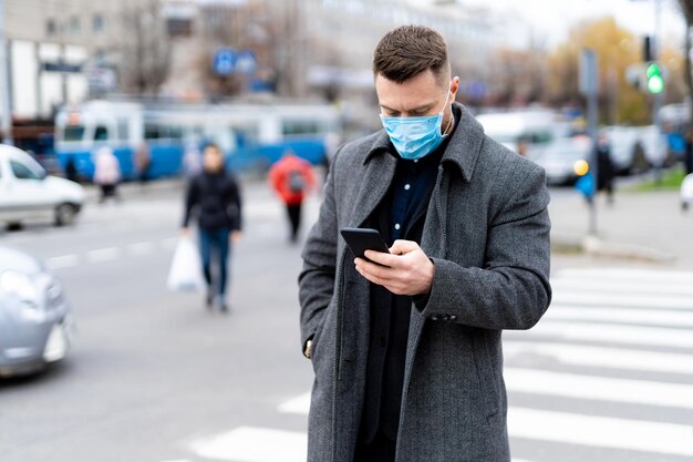 交差点の近くに立って電話で検索しているマスクの思慮深い男。世界でのコロナウイルスのパンデミック。