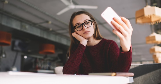 Thoughtfully female student holding mobile phone and taking selfie photos with front camera