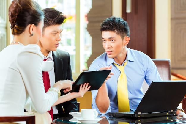 Three Asian Chinese office people or businessmen and businesswoman having a business meeting in a hotel lobby discussing documents on a tablet computer while drinking coffee