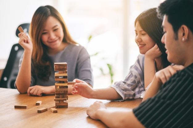 Three asian friends sitting and playing tumble tower wooden\
block game together with feeling happy