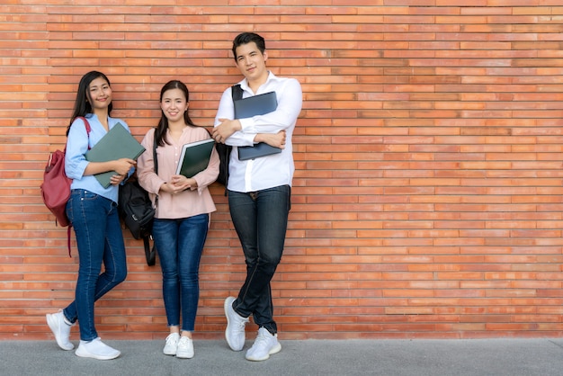 Photo three asian smiling student holding book and laptop posing on brick wall in campus.