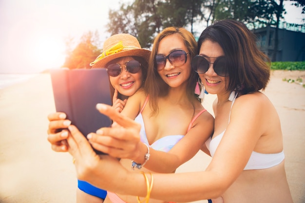 夏のビーチでリラックスして、スマートフォンでselfieを取って3つのアジアの女性