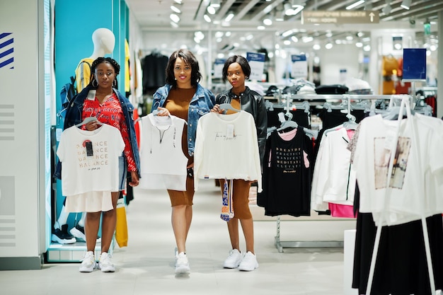 Three attractive african american woman in brown tunic dress posed at clothes store and hold shirts on hangers It's time for shopping