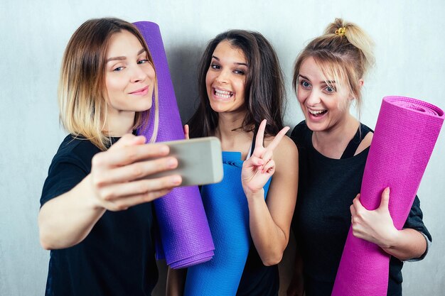 Three attractive and young female girlfriends hold a roll yoga\
mat and do selfie on the phone concept of yoga practice and female\
friendship