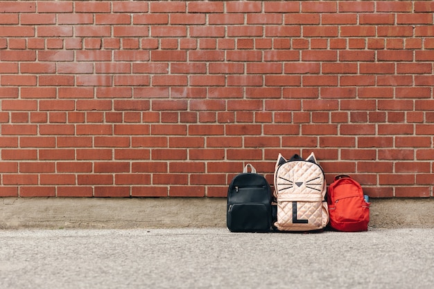 Three backpacks, black, pink, red, in front of the red brick wall