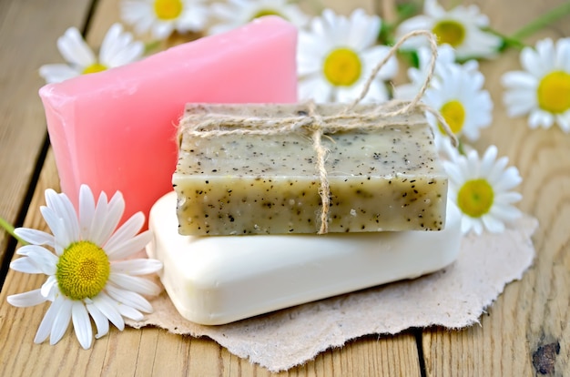 Photo three bars of soap pink, brown and white on a piece of paper, daisy flowers on a background of wooden boards