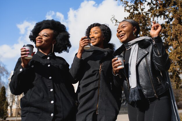 Photo three beautiful dark-skinned girls with long hair standing in a city and drinking a coffee