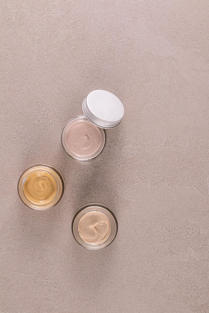 Foto tre bellissimi barattoli di vetro per i cosmetici con una delicata consistenza di crema per la cura del viso e del corpo su uno sfondo beige di pietra