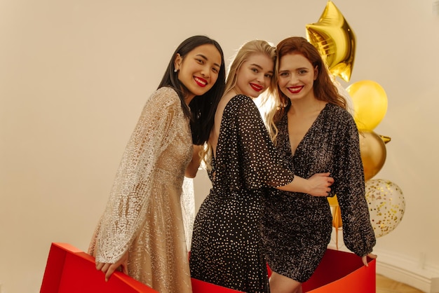 Three beautiful young interracial girls in sparkly dresses celebrating Women's Day smiling standing in big box Weekend concept