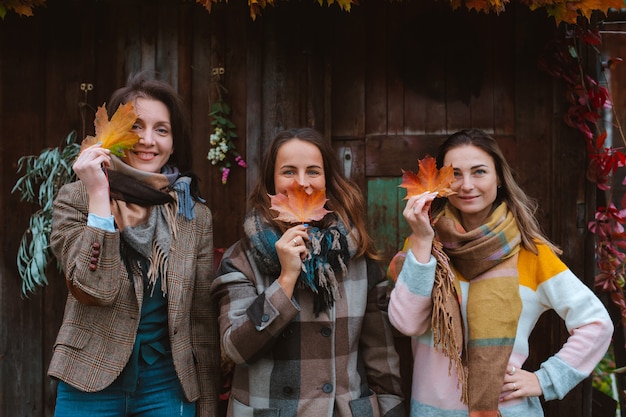 Three beautiful young women, covering their faces with a yellow autumn leaf, smiling on an old wooden background. The autumn fashion season.