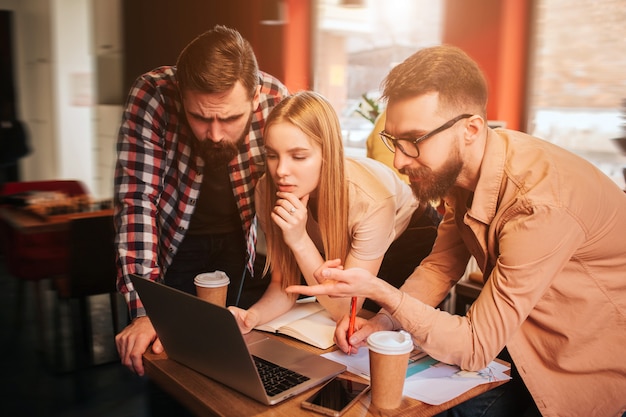 Three best friends are standing near table with computer on it and looking to the screen. One of them is pointing on the screen. The other two are listening to him carefully.