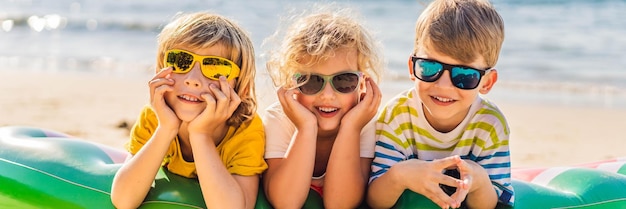 Foto tre bambini biondi in occhiali da sole sono sdraiati sul formato lungo della bandiera della spiaggia