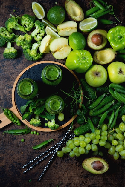 Three bottles of the most useful green smoothie from vegetables and fruits stand on a wooden wall in the center, and around green vegetables and fruits. Vertical photo. Top view.