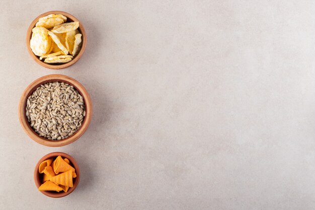 Three bowls of chips, crackers and sunflower seeds on stone surface.