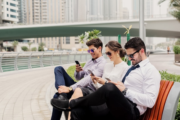 Three business people in suit taking a break from hard work . Sitting outside in Dubai Marina and having fun surfing on internet.