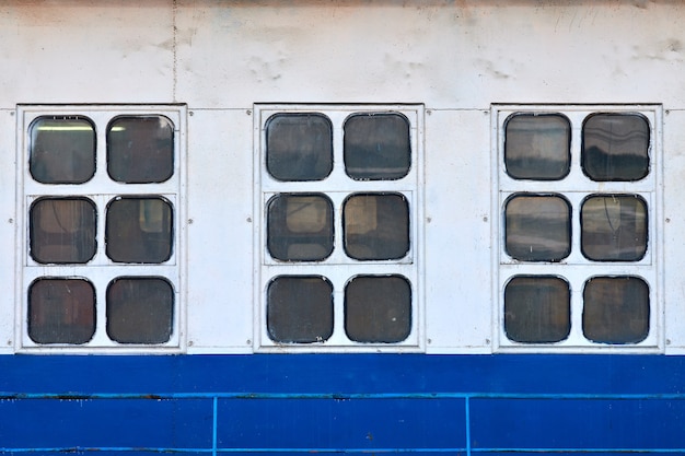 Three cabin windows and portholes on outboard side of ship. Close up of hull of vintage ocean liner. Old ship cabin windows, white and blue wall.