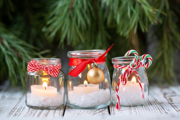 Three candles in glass jars with fir on holiday background Cozy handmade holiday home decor glass jar with candle decorated with red ribbon Christmas decorations