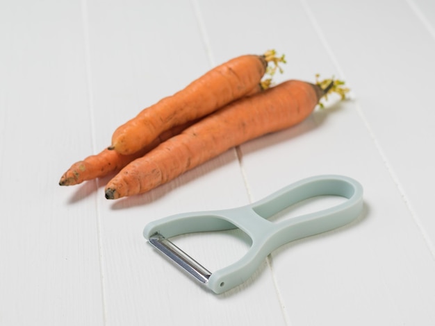 Три моркови и овощечистка на белом деревенском столе. Чистка моркови специальным ножом.