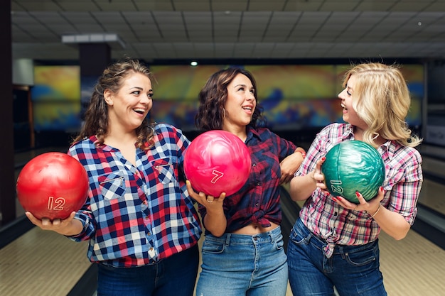 Three cheerful attractive girlfriends hold bowling balls and look at each other.