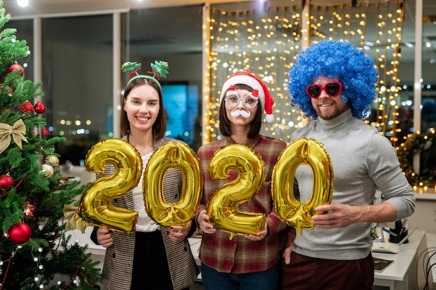 Three cheerful colleagues holding inflatable numbers while standing by decorated Christmas tree in office