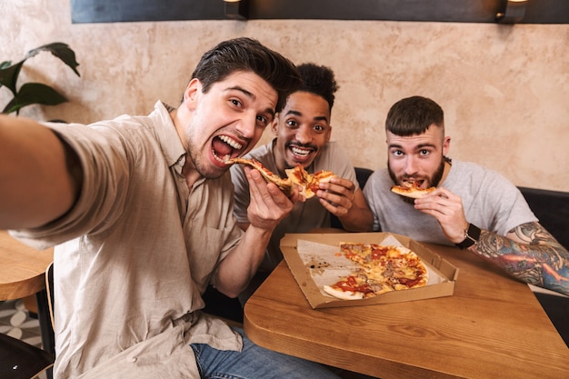 Three cheerful men eating pizza at the cafe table indoors, taking a selfie