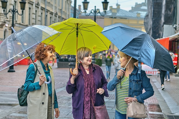 Три веселые женщины средних лет с разноцветными зонтиками гуляют по центру города во время дождя.