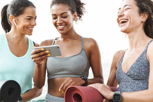 Three cheerful young sport girls standing at the beach, holding fitness mats, using mobile phone