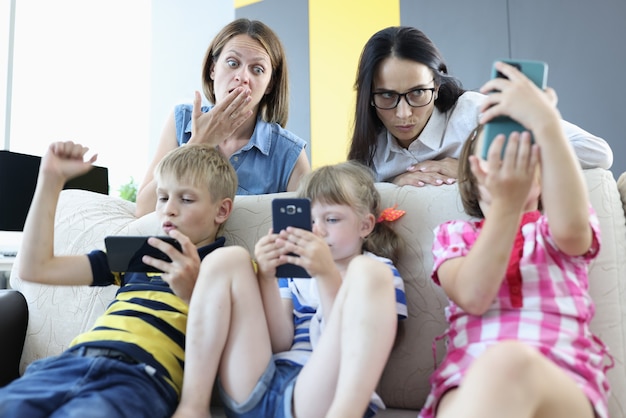 Фото Трое детей сидят на диване со смартфонами в руках, играют в онлайн-игры со спины дивана, две женщины стоят и испуганно смотрят на экран телефона.