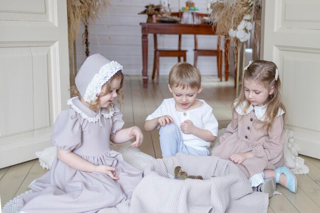 Фото Трое детей маленькие блондинки в старинных платьях и милый маленький мальчик в гостиной в загородном доме в весенний день играют с маленькими утками утками счастливое детство простая уютная жизнь счастливая пасха