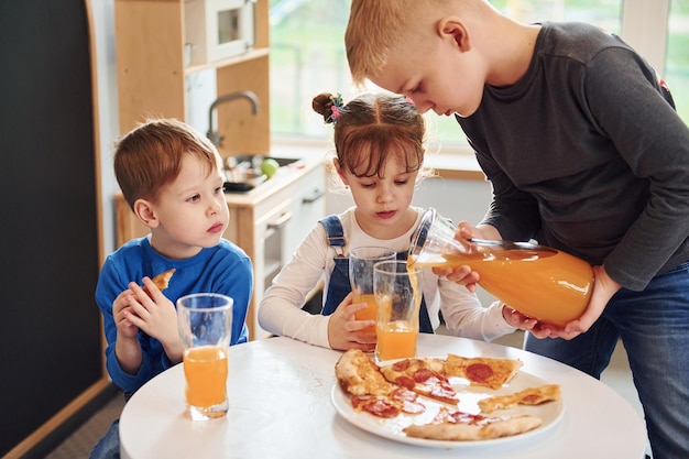 Three children sitting indoors by the table and eating pizza with orange juice together