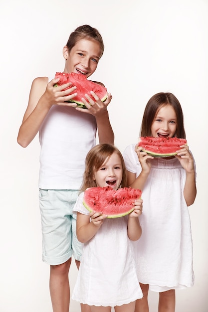 Three children in the studio stand on  white background and eat pieces of watermelon and laugh Brother two sisters In bright clothes barefoot Happy childhood pampering Proper power supply