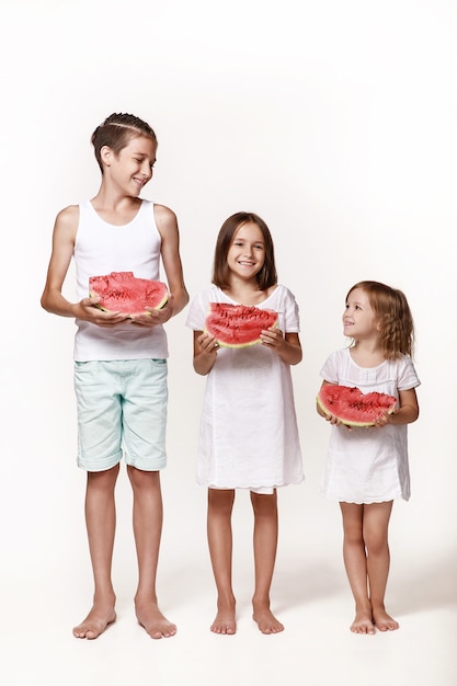 Three children in the studio stand on a white background and hold pieces of watermelon Brother two sisters In bright clothes barefoot Happy childhood Proper power supply