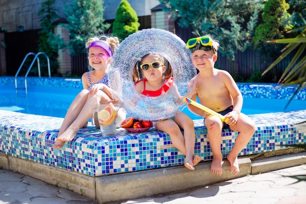 여름에 세 아이가 선글라스를 쓰고 수영장 옆에 앉아 레모네이드를 마신다.