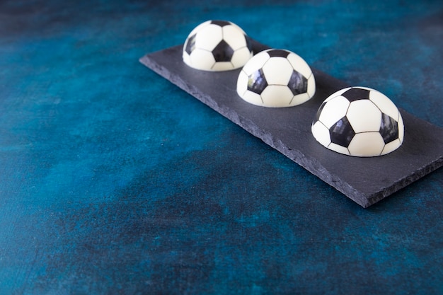 Фото Три шоколадных торта в форме футбольных мячей на прямоугольной черной тарелке в углу с копией пространства. креативный подарок фанату европейского футбола