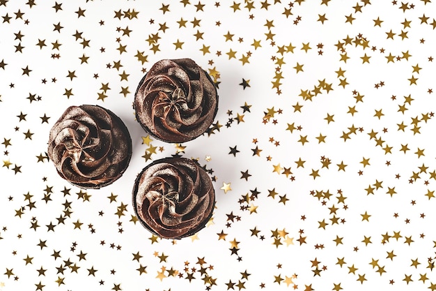 Three chocolate cupcakes with chocolate icing on white background with golden star shaped confetti