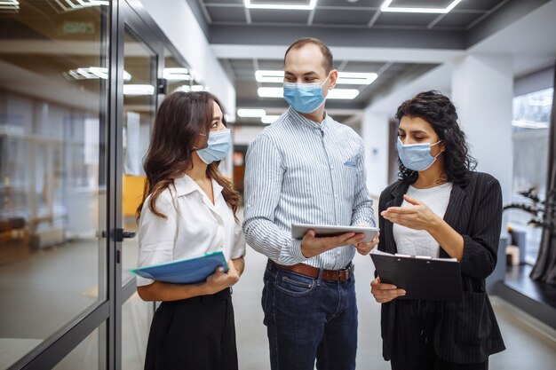 Three colleagues wearing medical masks discuss business at office corridor during coronavirus pandemic quarantine.