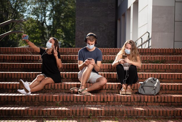 Three college students sitting on the stairs and using their smartphones wearing a face mask