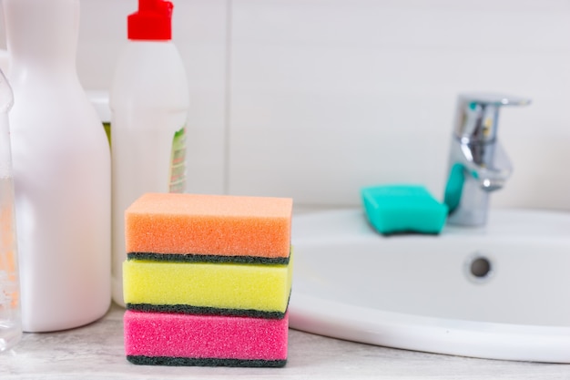 Tre spugne colorate per sgrassare e pulire la casa impilate l'una sull'altra accanto a un lavabo da bagno bianco pulito con un flacone spray per detersivo