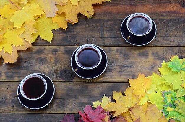 A three cups of tea among a set of yellowing fallen autumn leaves on a background surface of natural wooden boards of dark brown color